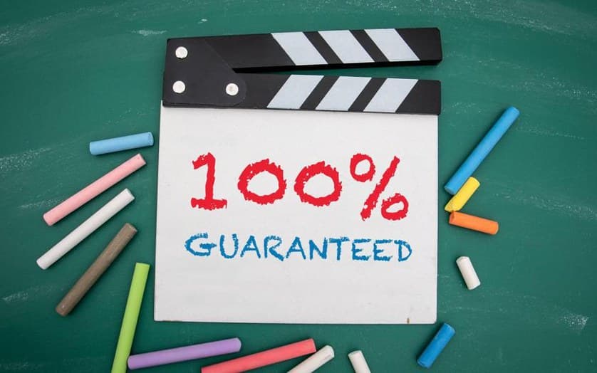 Filmklappe mit der Aufschrift „100% Guaranteed“ auf grünem Hintergrund und bunten Kreidestücken