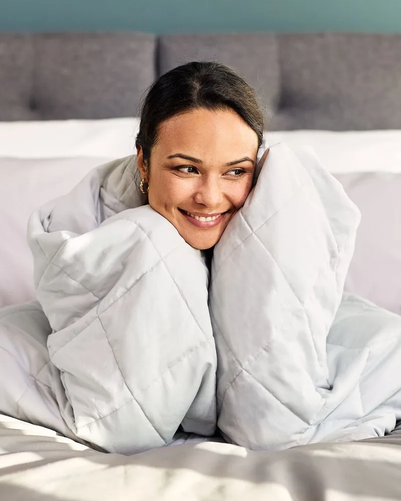 Lächelnde Frau im Bett, eingekuschelt in eine hellblaue Emma-Gewichtsdecke